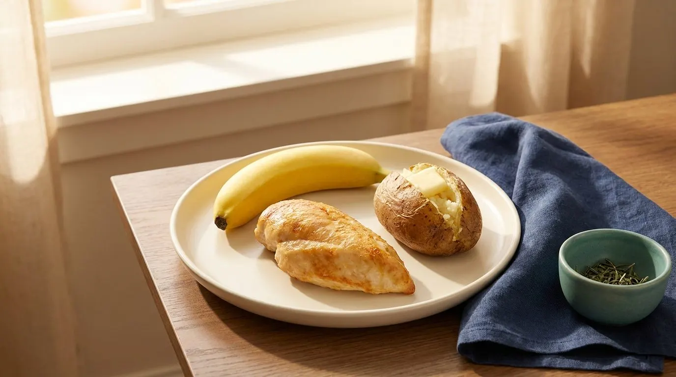 A cooked chicken breast, a ripe banana, and a baked potato arranged together on a simple plate with warm natural lighting