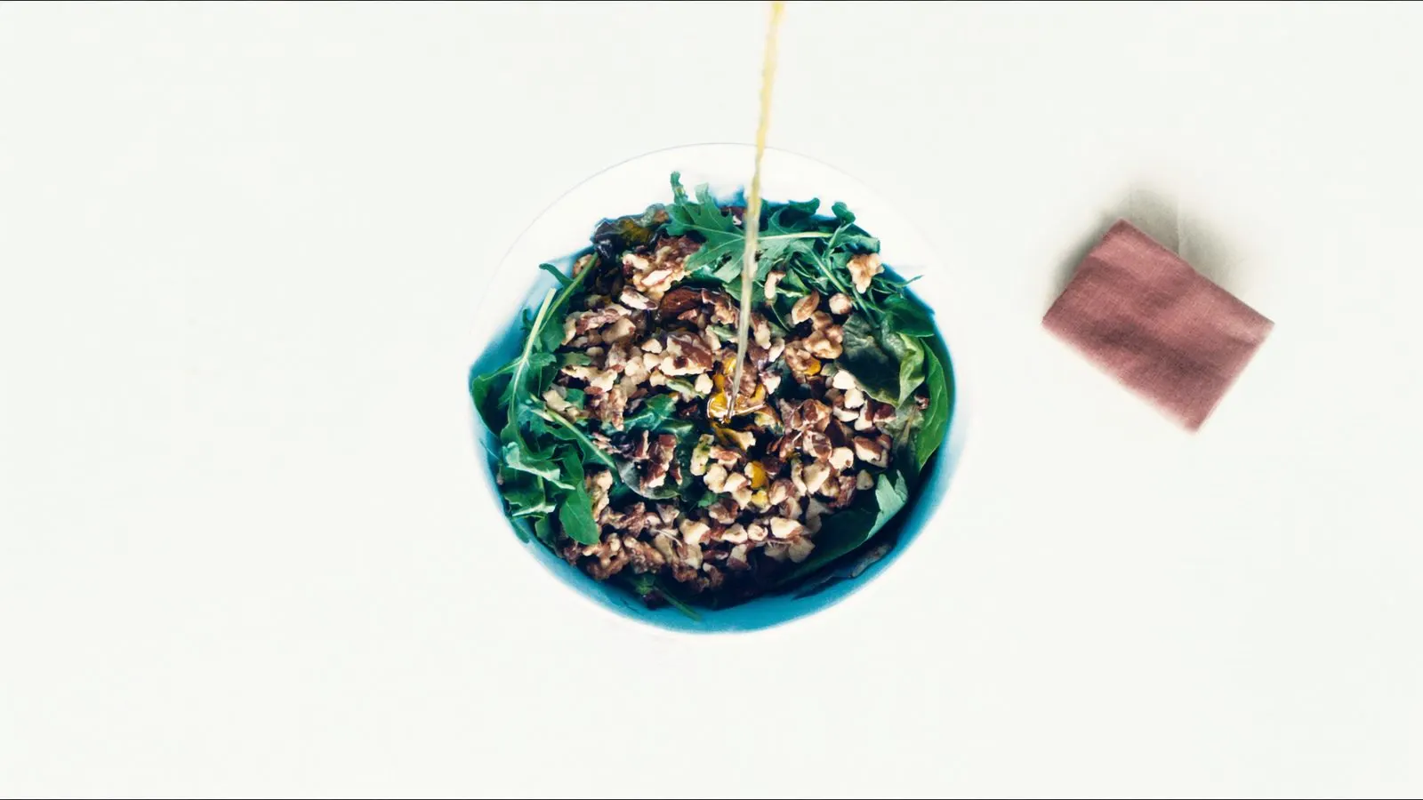 Editorial overhead of a large salad bowl with visible oil drizzle and nuts on cream linen, single dusty rose napkin.