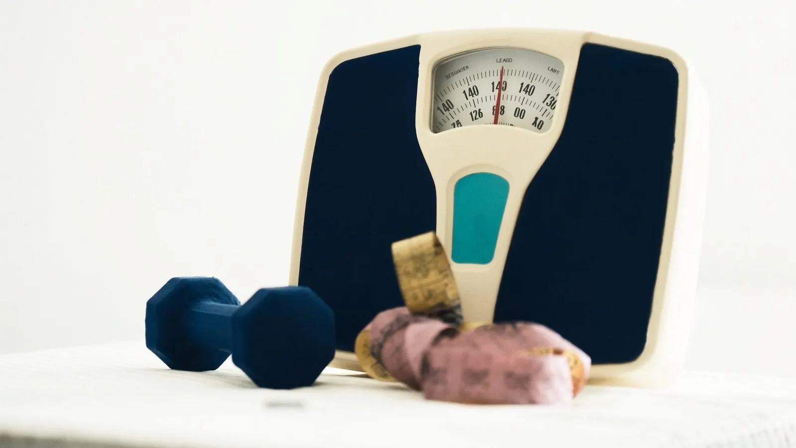 Editorial close-up of a bathroom scale next to a measuring tape and a small dumbbell on cream linen.