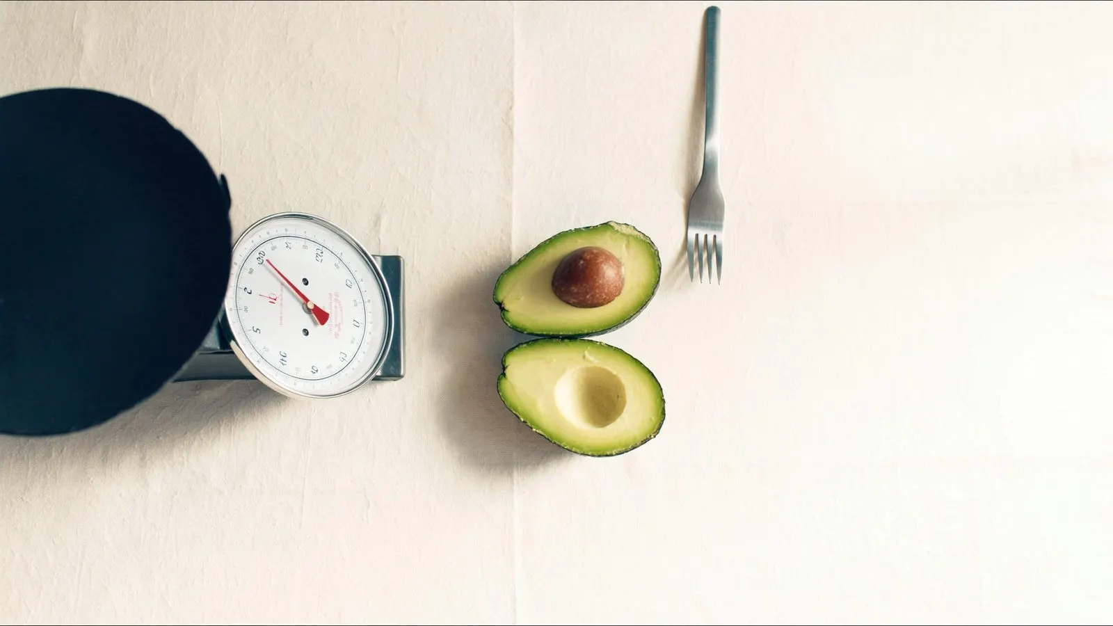 Editorial top-down of a kitchen scale with a single avocado and a fork on cream linen, soft daylight.