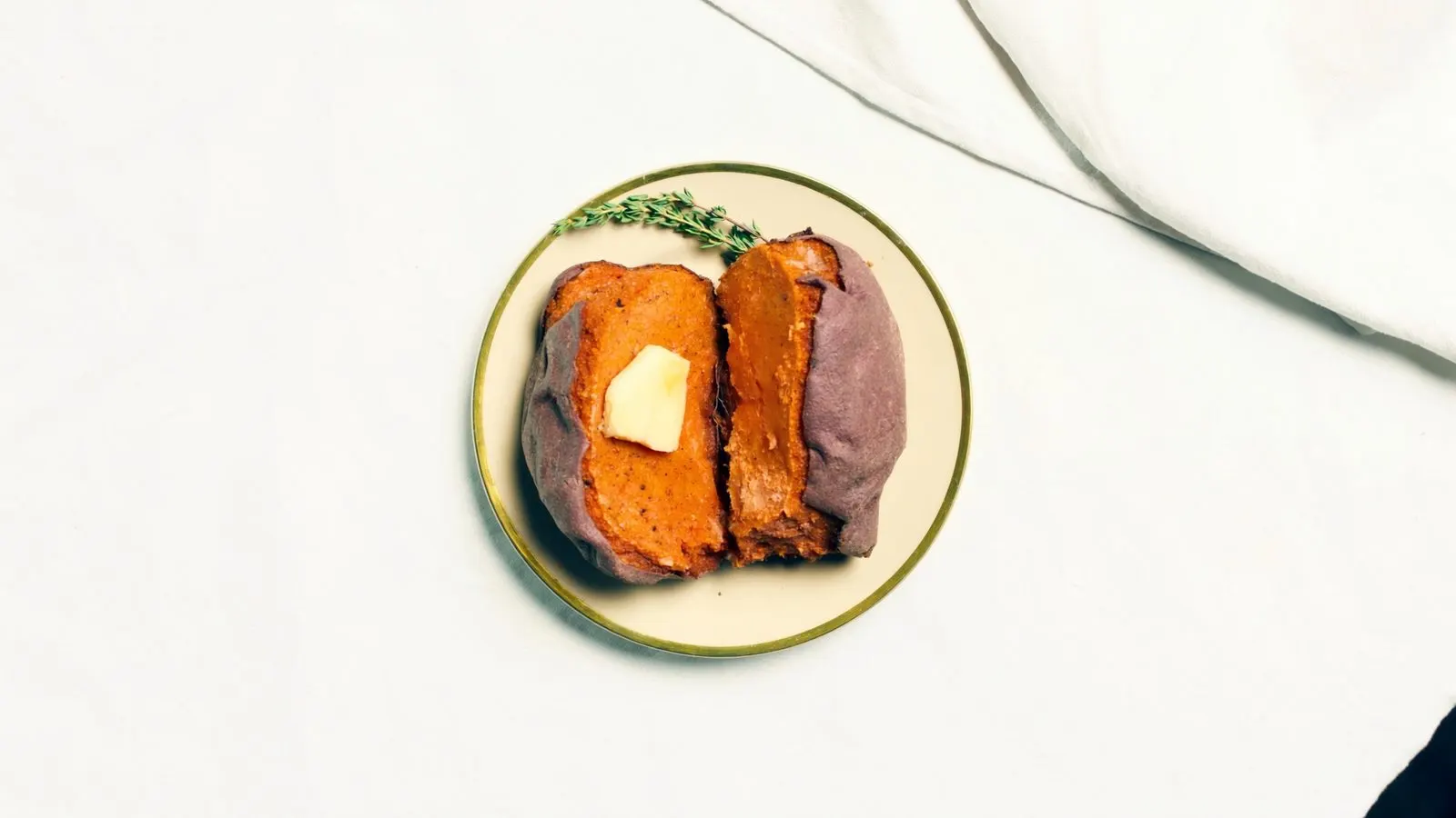 Editorial close-up of a baked sweet potato split open with a single pat of butter melting, cream linen background.