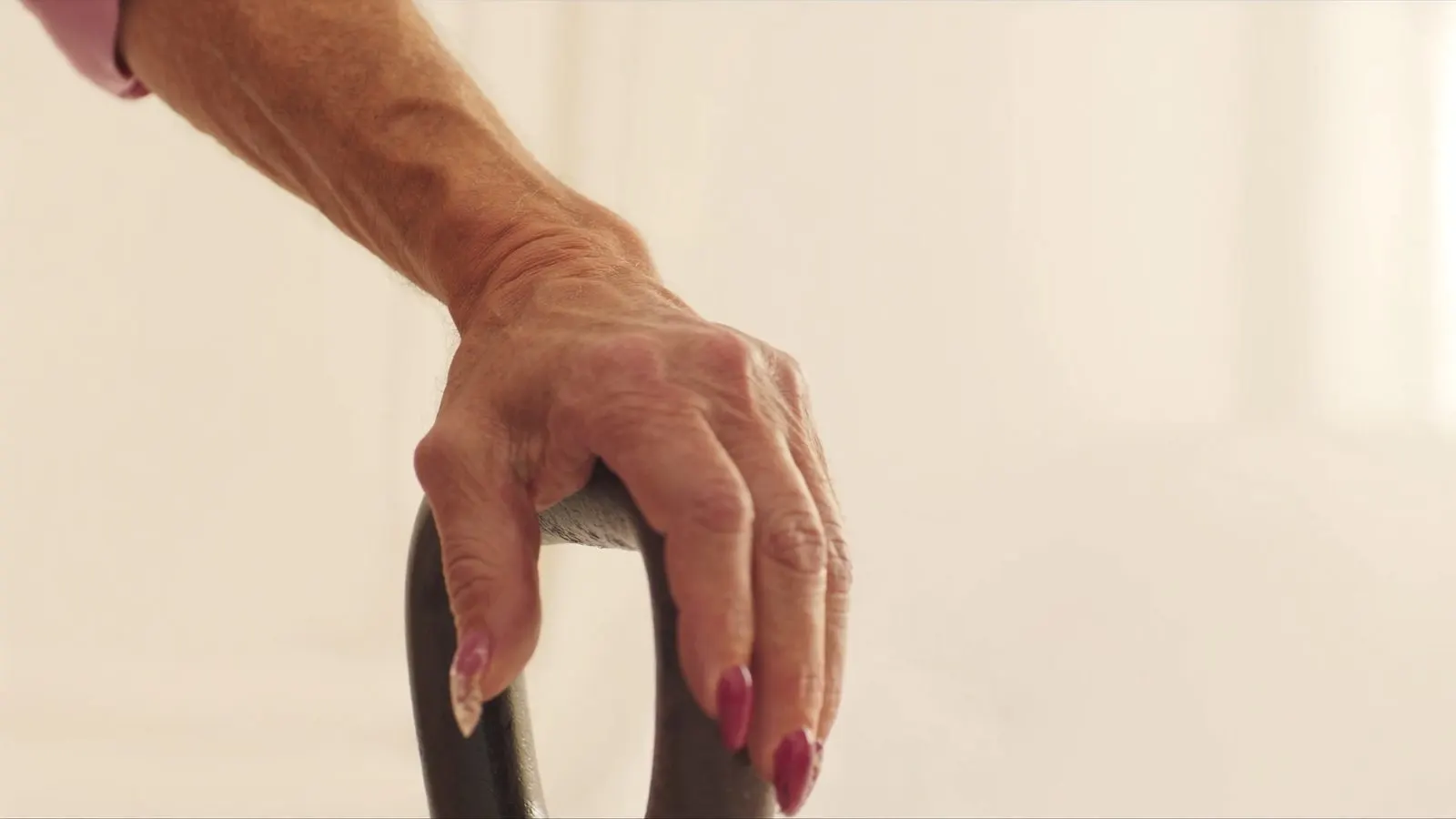Editorial close-up of a mature woman's hand resting on a kettlebell handle, cream and dusty rose palette.