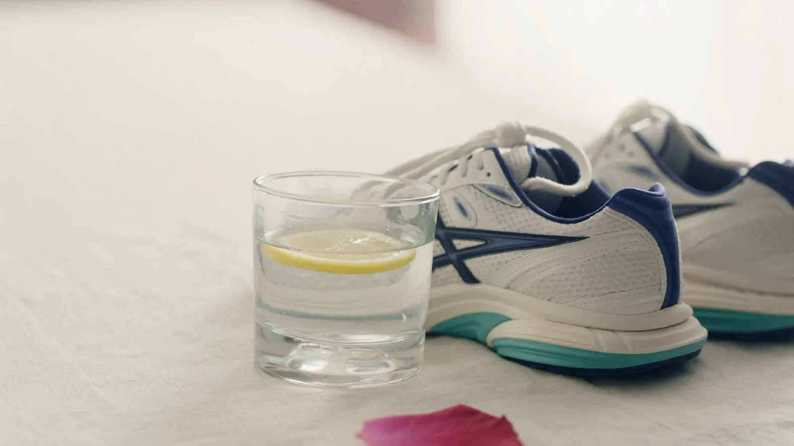 Editorial still life of a glass of water with a single lemon slice next to premium running shoes on cream linen surface.