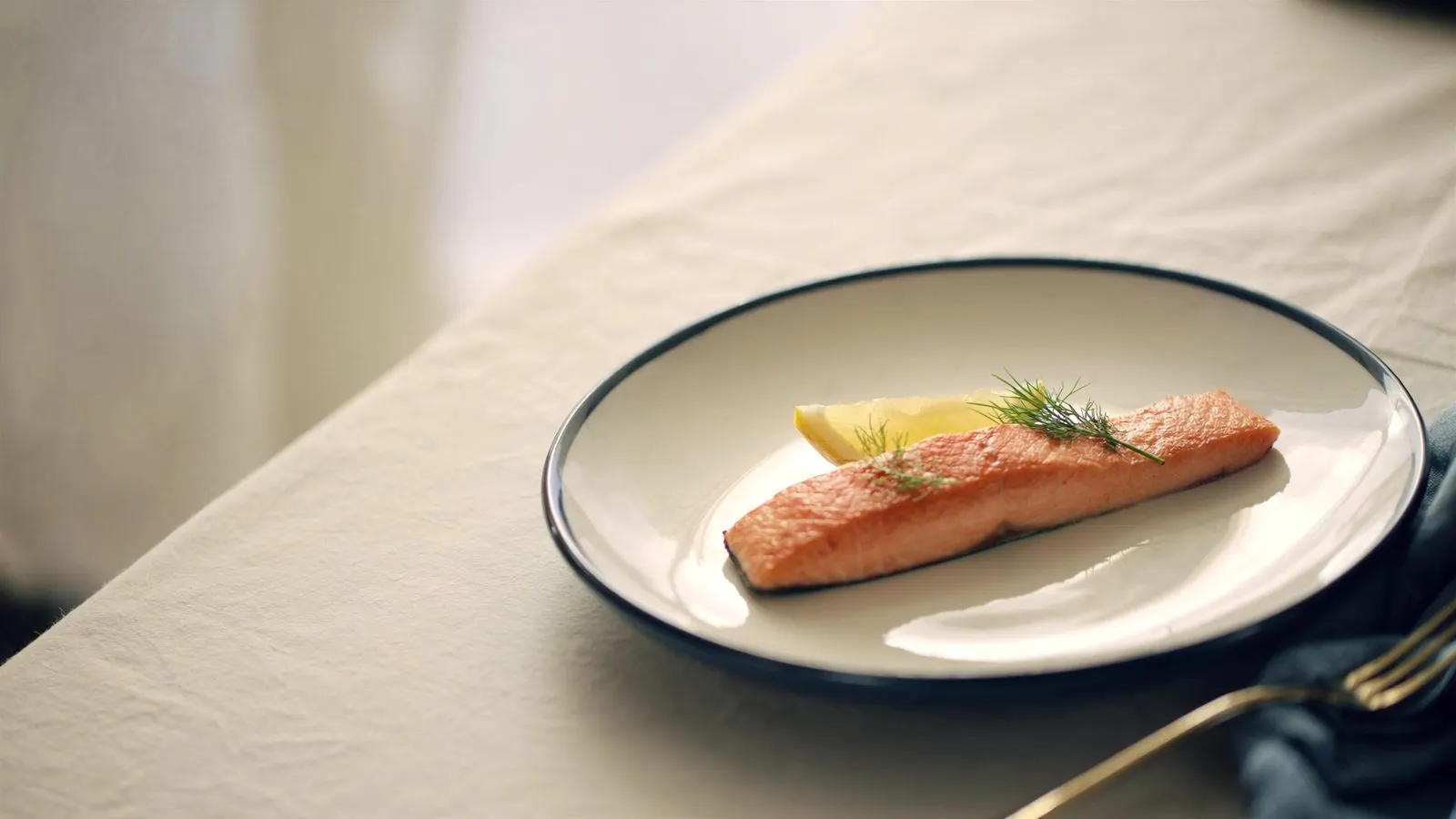 Editorial close-up of a small portion of grilled salmon with a wedge of lemon on cream porcelain, soft window light.