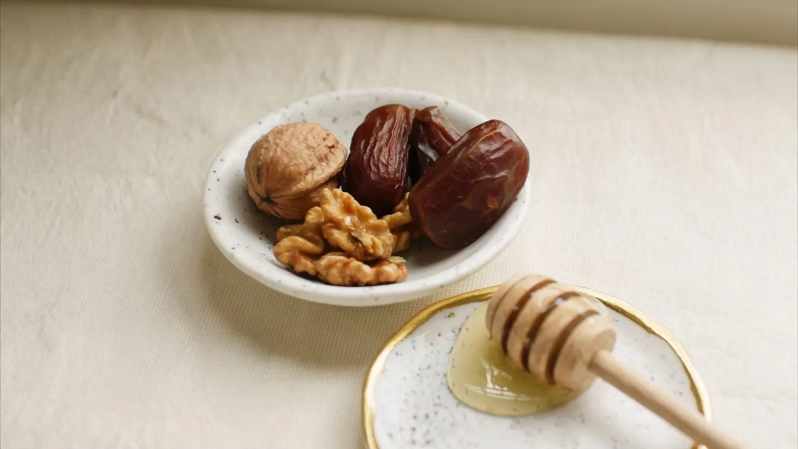 Editorial close-up of a small porcelain dish of dried dates and walnuts beside a wooden honey dipper, cream linen, soft daylight.