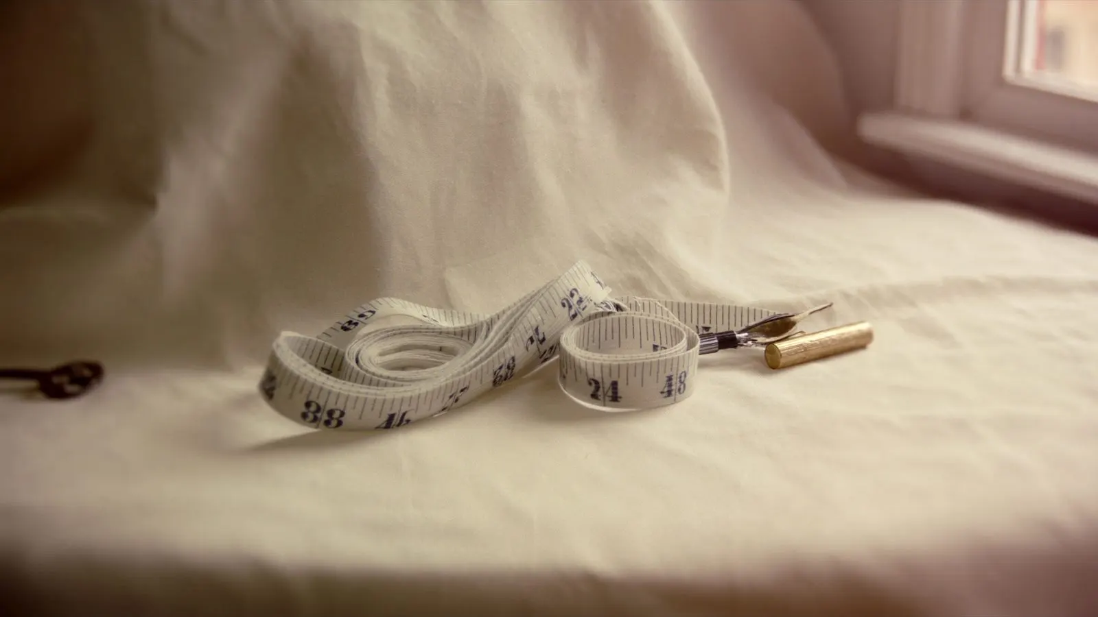 Editorial close-up of a measuring tape coiled neatly on cream linen with a small brass pen, soft daylight.